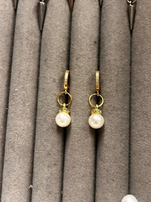 Guldörhängen med pärlor och stenar - Snygga örhängen i guldfärg med vita pärlor och små gnistrande stenar på bygeln. Hängande design som ger en elegant look. Perfekta för dig som vill ha något klassiskt men ändå trendigt. Materialet ser ut att vara metall med pärlimitation.