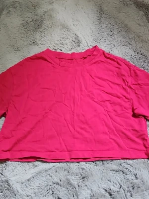 Croppad rosa t-shirt - En enkel croppad t-shirt i stark rosa färg. T-shirten har korta ärmar och rund halsringning. Perfekt för dig som gillar en avslappnad och trendig stil. Materialet känns mjukt och skönt, troligtvis bomull.
