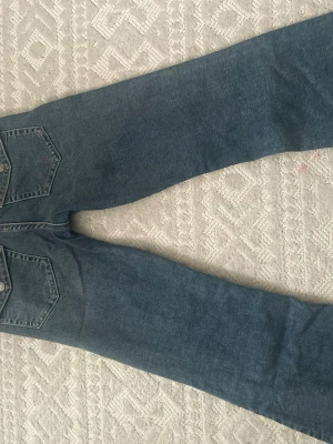 Blå raka jeans med bakfickor - Klassiska blå jeans med raka ben och två stora bakfickor med knappdetaljer. Jeansen har hög midja och är tillverkade i ett robust denimtyg. Perfekta för en avslappnad och trendig look.