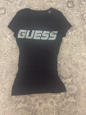 Svart GUESS t-shirt med strass - Snygg svart t-shirt från GUESS med stor logga framtill i glittrande strass. Klassisk rund halsringning och korta ärmar. Perfekt för dig som vill sticka ut lite extra med en enkel men ändå lyxig look.