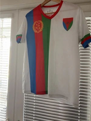 Eritrea t-shirt med flaggmotiv - Snygg t-shirt med Eritreas flagga och emblem på bröstet samt texten 'ERITREA' på ryggen. T-shirten har korta ärmar och är vit med blå, röd och grön rand på framsidan. Röd krage och detaljer på ärmarna. Perfekt för dig som vill visa stolthet för Eritrea.