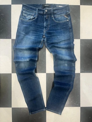 Blå slim fit jeans från Replay - Snygga blå slim fit jeans från Replay med klassisk femficksmodell och slitna detaljer. Jeansen har en tvättad look med ljusare partier och kontrastsömmar. Perfekta för en trendig och avslappnad stil.