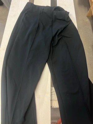 Svarta kostymbyxor från Gina Tricot - Säljer ett par svarta kostymbyxor från Gina Tricot. Byxorna har hög midja, pressveck och raka ben. Klassisk look med bälteshällor och diskret dragkedja. Materialet känns mjukt och följsamt, perfekt för en stilren outfit.