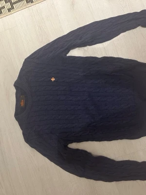 Mörkblå kabelstickad tröja från Morris  - Snygg mörkblå kabelstickad tröja med rund hals och ett litet broderat gult emblem på bröstet. Tröjan har långa ärmar och ribbade muddar vid ärmslut och nederkant. Perfekt för lager på lager och en klassisk look.