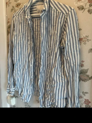 Randig blåvit linneskjorta med knappar - Säljer en långärmad skjorta med blå och vita ränder. Skjortan har klassisk krage och knäppning framtill. Tillverkad i ett lätt och luftigt material, perfekt för en avslappnad stil.