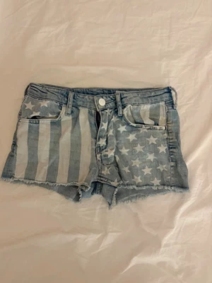 USA jeansshorts  - Snygga jeansshorts i storlek 146, men jag skulle säga att de även passar XXS eller en liten XS. Häftigt USA-inspirerat mönster med stjärnor och ränder – perfekta till sommaren.  Det finns en fläck, men den syns på en av bilderna. I övrigt är shortsen i fint begagnat skick.  Hör gärna av dig om du har frågor! 🌸