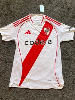 River Plate matchtröja  - Snygg vit River Plate matchtröja från Adidas med röd diagonal rand och klubbmärke på bröstet. Tröjan har röda detaljer på axlarna, HEAT.RDY-material och slim fit-passform. Perfekt för fotbollsträning eller att bära på läktaren.