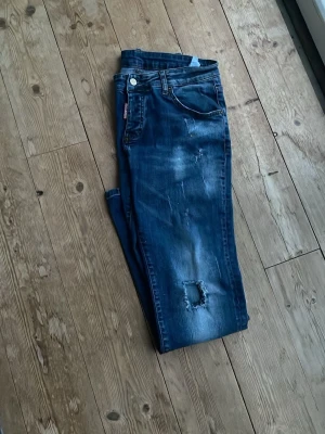 Blå slitna jeans med patchdetalj - Säljer ett par blå jeans med slitningar och patchdetalj på ena benet. Jeansen har klassisk femficksmodell, normal midja och dragkedjegylf. Snygg tvätt med lätt urtvättade partier och en avslappnad look.