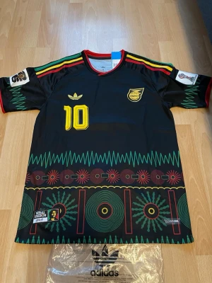 Jamaica fotbollströja Adidas Marley ( CUSTOM ) - Snygg svart Jamaica fotbollströja från Adidas med färgstarka detaljer i grönt, rött och gult. Nummer 10 och Marley på ryggen, samt Reggae Boyz-tryck. Unik design med grafiska mönster och klubbmärke på bröstet. Perfekt för dig som gillar fotboll och reggae. Har world cup patches. Support för landet som inte fick spela detta år.