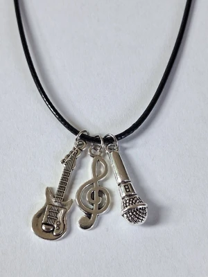 Halsband med musikinspirerade berlocker - Svart vaxade läderhalsband med tre silvriga berlocker i form av en elgitarr, en g-klav och en mikrofon. Kedjan är tunn men slitstark och smidig, och berlockerna är detaljerade i metall. Perfekt för dig som älskar musik och vill ha en unik accessoar. Materialet rostfritt stål 
