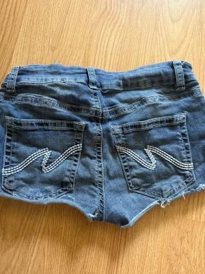 Blå denimshorts med dubbla knappar - Säljer ett par blå denimshorts från Nelly med dubbla knappar framtill och coola detaljer på bakfickorna. Shortsen har råa kanter och klassisk femficksdesign. Perfekta för varma dagar och en avslappnad stil. Dom sitter perfekt runt midjan, inte för löst men inte för tajt. Säljer på grund av för lite användning.