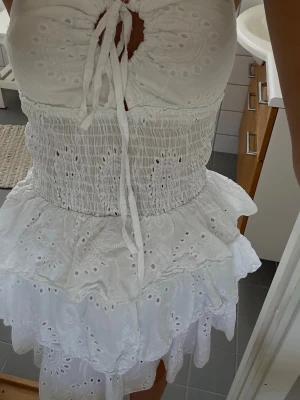 Vit omlottklänning med volanger - Söt vit kortklänning med broderade detaljer, omlottdesign och snörning framtill. Klänningen har volanger i flera lager och smockad midja för en bekväm passform. Perfekt för sommarens alla tillfällen.