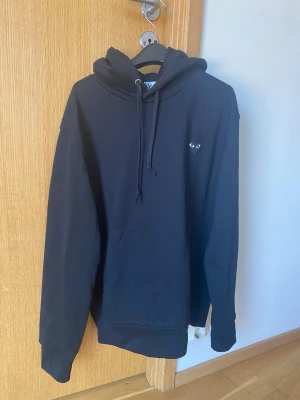 Svart hoodie med hjärtdetalj - Säljer en svart hoodie med huva och dragsko. På bröstet finns en liten hjärtformad patch med ögon, vilket ger en unik touch. Hoodien har långa ärmar, ribbade muddar och en klassisk passform. Perfekt för dig som gillar stilrena plagg med en twist.