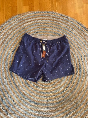 Mörkblå prickiga från Loro Piana - Snygga mörkblå badbyxor från Loro Piana med vita små prickar och vit dragsko i midjan. De har elastisk midja och innerbyxa i beige. Perfekta för stranden eller poolen och ger en klassisk men ungdomlig look. Helt nya med tags, nypris €490. Passar en L, väldigt små i storlek.