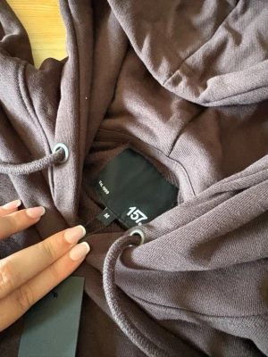 Brun hoodie från 157 med huva oanvänd - Mysig brun hoodie från 157 med klassisk känguruficka och justerbar huva med dragsko. Tillverkad i mjukt bomullsliknande material och har långa ärmar. Perfekt för en avslappnad och chill stil.