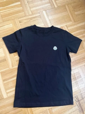 Svart t-shirt med liten logga - En stilren svart t-shirt i bomull med rund hals och korta ärmar. På bröstet sitter en liten broderad logga som ger en clean och modern look. Perfekt till jeans eller shorts för en enkel men snygg outfit. Storlek S helt ny