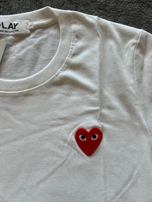 Comme des Garçons t-shirt - Vit t-shirt från Comme des Garçons PLAY med det ikoniska röda hjärtat med ögon broderat på bröstet. Klassisk rund hals och korta ärmar. Tillverkad i mjuk bomull som känns skön mot huden. Perfekt för en clean och stilren look. Storlek S