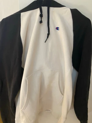 Champion hoodie svart/vit - Snygg hoodie från Champion med svart huva och ärmar samt vit kropp. Klassisk broderad Champion-logga på bröstet. Hoodien har känguruficka framtill och justerbar dragsko i huvan. Perfekt för en avslappnad streetwear-look.