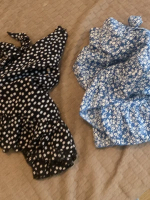 Två korta sommarklänningar mönstrade säljer två för 30kr - Säljer två korta klänningar, en svart med vita prickar och en blå med vitt blommönster. Båda har volangdetaljer och omlottknytning i midjan. Perfekta för varma dagar och ger en söt och trendig look.