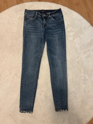 Blå skinny jeans med pärldetaljer - Snygga blå skinny jeans med dekorativa pärlor och rosetter längst ner vid bensluten. Jeansen har klassisk femficksmodell och normalhög midja. Perfekta för dig som vill ha något extra på dina jeans!