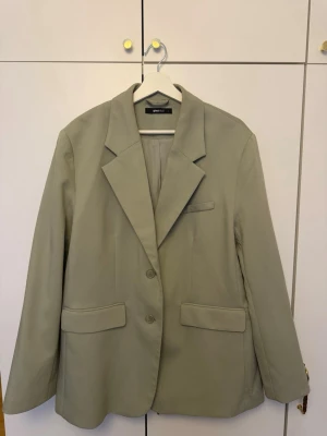 Beige kavaj från Gina Tricot - Stilren beige kavaj från Gina Tricot med klassisk passform, två knappar framtill och tre fickor. Kavajen har breda slag och är helfodrad för en snygg siluett. Perfekt för dig som vill ha en enkel men trendig look.