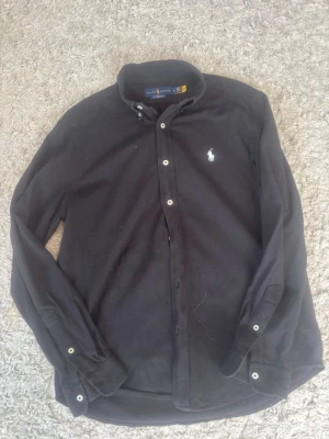 Svart skjorta från Ralph Lauren - Klassisk svart skjorta från Ralph Lauren med button-down krage och vita knappar. Skjortan har lång ärm och den ikoniska broderade loggan på bröstet. Tillverkad i mjuk bomull som känns skön mot huden. Perfekt för en stilren och avslappnad look. Storlek L på skjortan men sitter som M