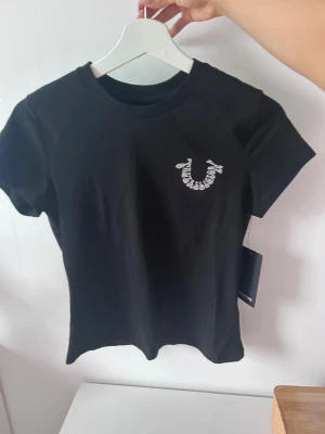Svart True Religion t-shirt - Snygg svart t-shirt från True Religion med ikoniskt logotryck i vitt framtill och stort tryck på ryggen. T-shirten har rund hals, korta ärmar och är gjord i mjuk bomull. Perfekt till jeans eller shorts för en clean och trendig look.