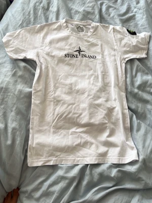 Vit Stone Island t-shirt - Vit t-shirt från Stone Island med klassisk logga tryckt på bröstet och ikonisk patch på ärmen. T-shirten har rund hals och korta ärmar. Perfekt basic-plagg med clean look och streetwear-vibe.