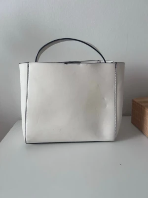 Vit stilren handväska i konstläder - Snygg vit handväska i konstläder med minimalistisk design och raka linjer. Väskan har ett topphandtag och ett rymligt huvudfack med innerficka med dragkedja. Svarta kontrastsömmar ger en modern touch. Perfekt för dig som gillar enkel och stilren look.