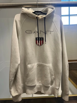 Vit hoodie från GANT med logga - Säljer en stilren vit hoodie från GANT med broderad logga och sköld på bröstet. Hoodien har svart snörning i huvan och en stor magficka. Perfekt för dig som gillar klassisk och enkel stil.