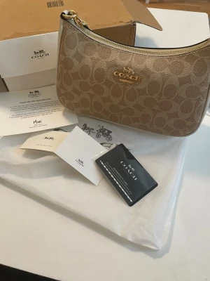 Beige axelremsväska från Coach - Snygg beige handväska från Coach med klassiskt monogrammönster och guldfärgade detaljer. Väskan har en avtagbar och justerbar axelrem i ljusbeige läder samt dragkedja upptill. Perfekt för dig som gillar stilrena accessoarer med lyxig känsla.