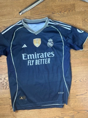 Mörkblå Real Madrid matchtröja - Säljer en mörkblå Real Madrid matchtröja från Adidas med vita detaljer och klubbmärke på bröstet. Tröjan har v-ringad krage, korta ärmar och tryck med 'Emirates Fly Better' på framsidan. Perfekt för fotbollsträning eller att bära som supporter. Priset är inte fast!
