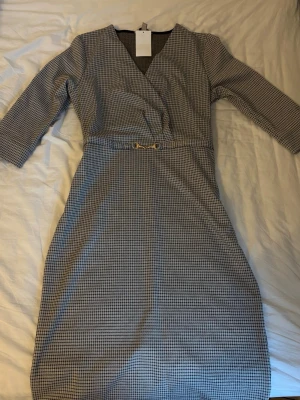Rutig omlottklänning  - Stilren rutig klänning från H&M med omlott-design och v-ringning. Klänningen har trekvartsärm och ett dekorativt guldfärgat spänne i midjan. Perfekt för dig som gillar klassiska mönster och en snygg siluett.