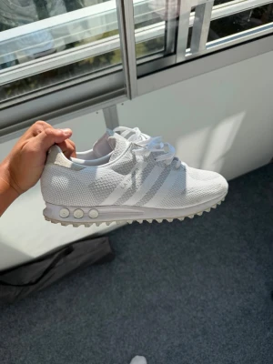 Adidas la trainers  - Snygga vita Adidas sneakers med stickad ovandel och klassiska tre ränder på sidan. Skorna har vita snören, låg profil och en unik sula med rundade detaljer. Perfekta för en clean och sportig look.