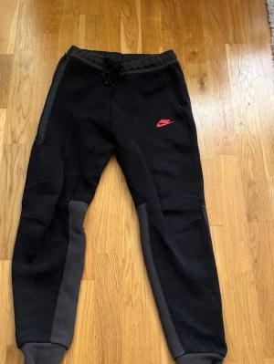 Svarta Nike mjukisbyxor med logga - Säljer ett par svarta mjukisbyxor från Nike med rosa logga på vänster lår. Byxorna har grå detaljer längs benen, dragsko i midjan och muddar vid bensluten. Perfekta för chill eller träning.