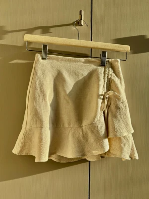 Beige omlottkjol med volang - Säljer en beige kort omlottkjol i linne med volangkant och knyt i sidan. Kjolen har en avslappnad och trendig look som passar perfekt till sommaren. Superfin att matcha med en enkel topp eller stickad tröja.