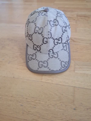 Beige keps med GG-mönster - Säljer en beige keps från Gucci med det klassiska GG-mönstret i brunt över hela kepsen. Kepsen har böjd skärm och bruna detaljer längs kanten. Perfekt för dig som vill ha en stilren och lyxig accessoar till din outfit.