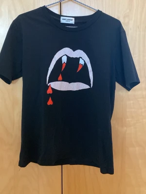 Saint Laurent Bloodluster T-shirt AW14 - Svart t-shirt från Saint Laurent med ett coolt tryck av vampyrtänder och röda blodsdroppar på bröstet. Klassisk rund hals och korta ärmar. Tillverkad i mjuk bomull som känns skön mot huden. Perfekt för dig som vill sticka ut med en edgy look.