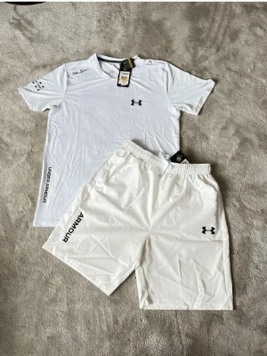 Vitt sommarset från Under Armour - Snyggt vitt sommarset från Under Armour med t-shirt och shorts. Båda plaggen har logga på bröst och ben samt tryckta detaljer. Tillverkade i lätt och snabbtorkande material, perfekt för träning eller sportaktiviteter. Elastisk midja på shortsen för skön passform.