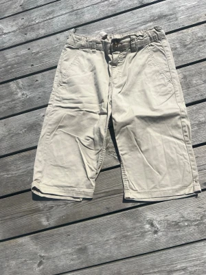 Beiga shorts från Kappahl - Säljer ett par beiga shorts från Kappahl i storlek 158. De har klassisk passform med fickor fram och bälteshällor i midjan. Perfekta för varma dagar och enkla att matcha med olika stilar.