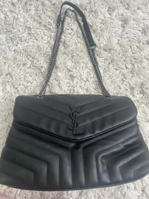 Svart YSL väska - Snygg svart quiltad handväska från YSL i mjukt läder med kedjerem och klassisk YSL-logga framtill. Väskan har lock med magnetlås och flera fack inuti, inklusive dragkedjefack. Perfekt för dig som vill ha en stilren och lyxig accessoar.