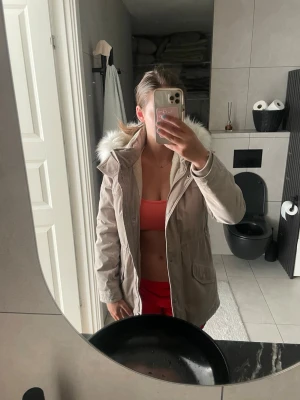 Beige parkas med fuskpäls - Snygg beige parkas med huva och fuskpälsdetalj. Jackan har dragkedja och knappar framtill samt två stora fickor. Insidan är fodrad med mjukt material för extra värme. Perfekt för kyliga dagar och enkel att matcha med olika outfits.