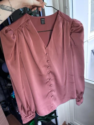 Långärmad blus i Koppar / rosa - Stilren kopparfärgad blus  i storlek XS. Blusen har v-ringning, klädda knappar framtill och puffiga axlar. Tillverkad i ett glansigt, mjukt material som ger en elegant look. Perfekt att matcha med jeans eller kjol för en trendig outfit.