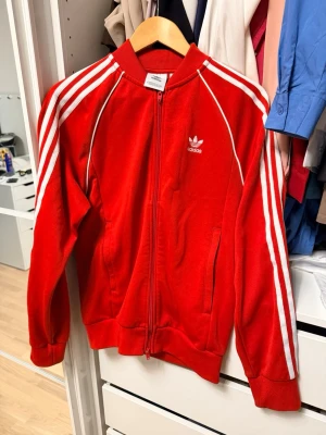 Röd Adidas track jacket med vita ränder - Klassisk röd Adidas track jacket med vita ränder längs ärmarna och Adidas-logga på bröstet. Dragkedja framtill, ribbade muddar och krage. Perfekt för en sportig och retro look. Tillverkad i mjukt material som känns bekvämt mot huden.