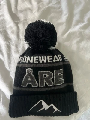 Svart mössa med tofs och Åre-tryck - Säljer en svart och grå stickad mössa med stor tofs på toppen. Mössan har texten 'ÅRE' och 'ONEWEAR' i vitt samt ett bergmotiv på den vikta kanten. Perfekt för kalla dagar och vintersäsongen.