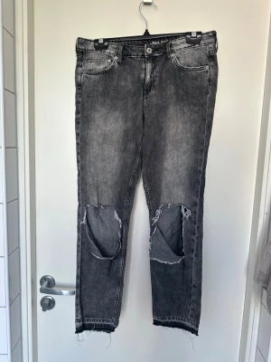 Grå slitna girlfriend jeans H&M storlek 27 - Säljer ett par gråa girlfriend jeans från H&M med låg midja och ankellängd. Jeansen har stora slitningar över knäna och råa, fransiga benslut. Tillverkade i denim med återvunna fibrer. Perfekta för en avslappnad och trendig look.