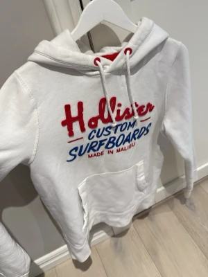 Vit hoodie från Hollister med tryck - Vit hoodie från Hollister med broderat rött och blått tryck på bröstet där det står 'Hollister Custom Surfboards Made in Malibu'. Hoodien har huva med dragsko och en stor magficka. Perfekt för en avslappnad och sportig stil.