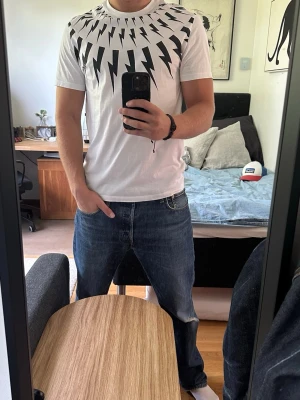 Neil Barrett Thunderbolt T-shirt - Säljer en vit Neil Barrett Thunderbolt t-shirt med ikoniskt svart blixtmönster runt halsen. T-shirten har korta ärmar, rund hals och är i slim fit. Tillverkad i mjuk bomull för skön känsla och stilren look.