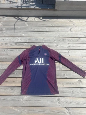 PSG (Nike) långärmad träningströja blå/röd - Säljer en långärmad träningströja från Paris Saint-Germain i mörkblått med röda detaljer och halv dragkedja. Tröjan har PSG-logga på bröstet och vit text 'Accor Live Limitless' framtill. Perfekt för fotbollsträning eller chill. Den är även från Nike 