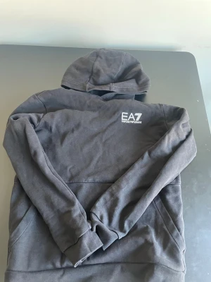 Svart hoodie från EA7 Emporio Armani - Snygg svart hoodie från EA7 Emporio Armani med vit logga på bröstet. Klassisk huva och magficka framtill. Tillverkad i mjukt bomullsmaterial som känns skönt mot huden. Perfekt för en avslappnad och trendig look.